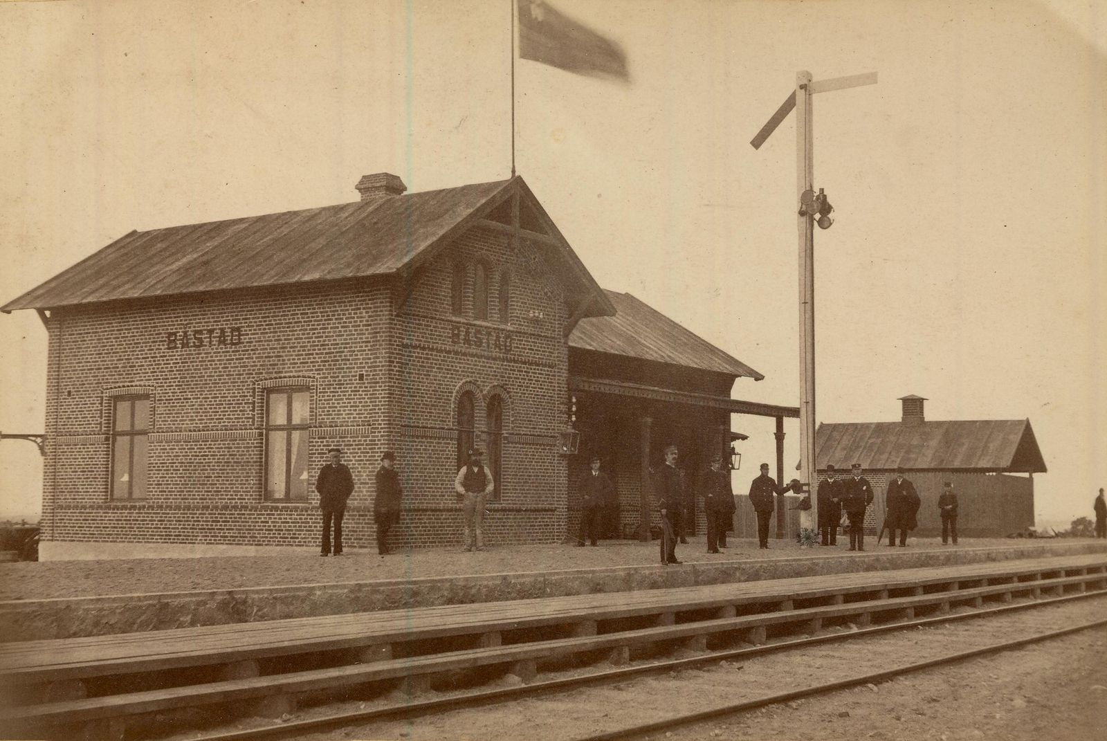 Fotografi av Båstads järnvägsstation, en tegelbyggnad med namnet BÅSTAD i formsten på två gavlar. Ett tiotal järnvägsmän står på perrongen, en semaforsignal reser sig till höger och spåret löper i förgrunden.