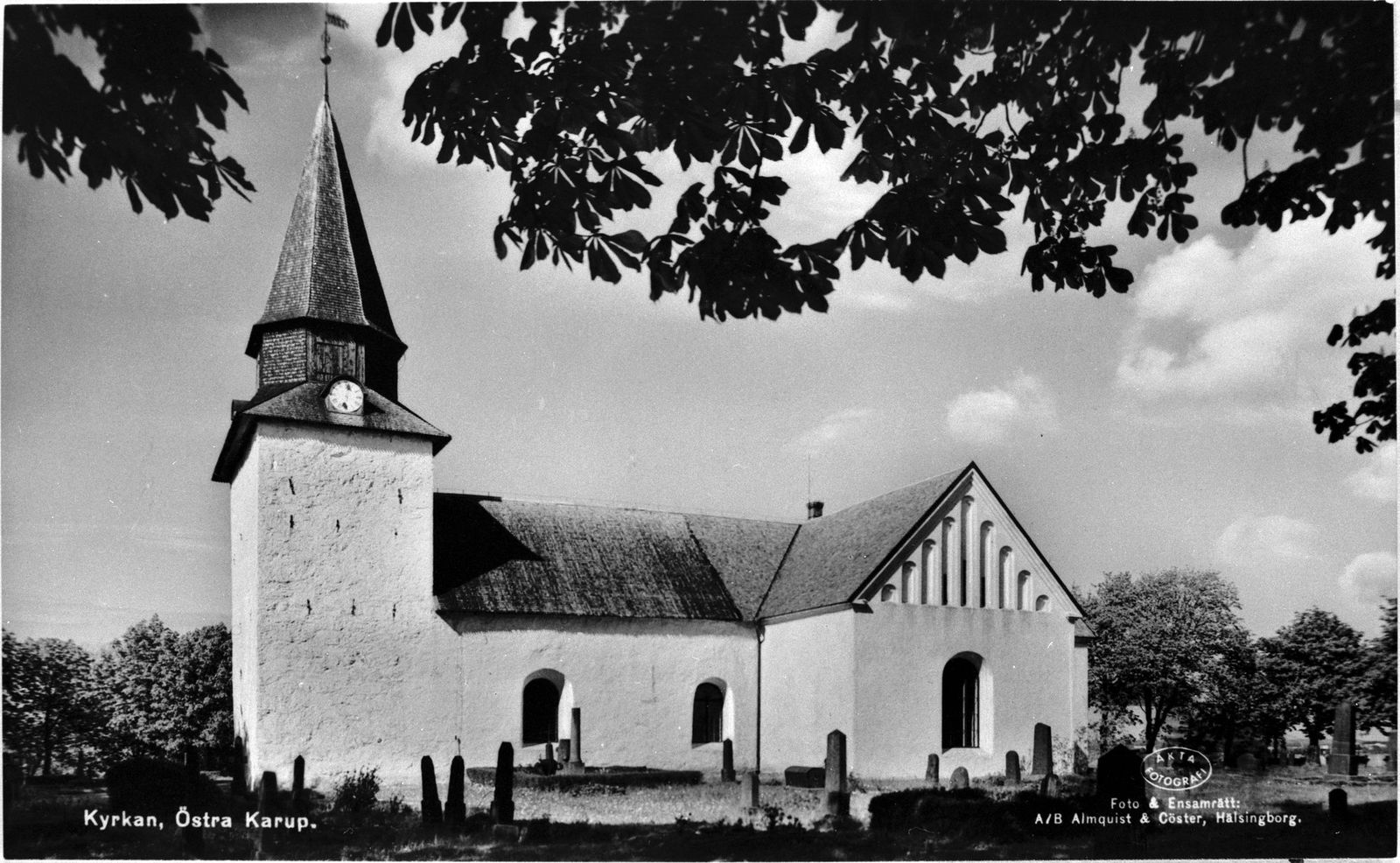Vykort som visar Östra Karups kyrka, en vitkalkad kyrka med torn och spira, med gravstenar på kyrkogården i förgrunden och kastanjelöv som hänger ner i bildens överkant.